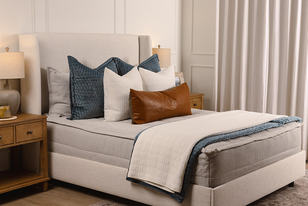 A bed made with light gray waffle texture zipper bedding and an assortment of neutral and velvet blue blankets and accessories. 