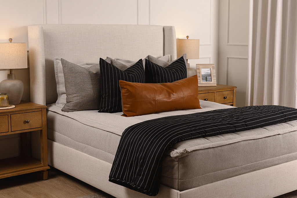 A bed made with light gray waffle textured zipper bedding and an assortment of tan, neutral, and black  throw pillows and blankets.