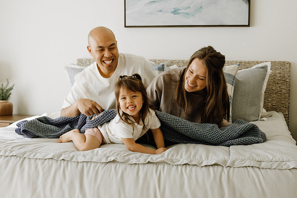 A mom, dad, and little girl laughing and cuddling in a blue and white stripe king bed with a blue, knitted blanket on top of them.