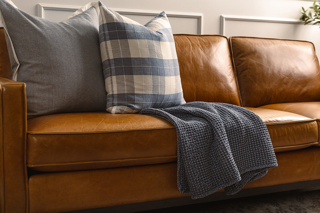 A blue plaid stitched pillow, a large blue pillow with white trim, and a dust blue waffle weave throw on an orange leather couch. 
