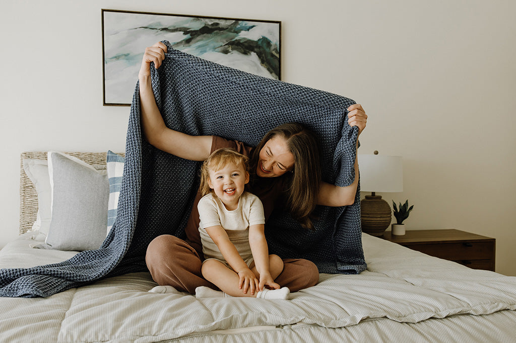 A mother and daughter sitting on a neutral bed with a dusty blue, waffle weave textured throw blanket wrapped around them. 
