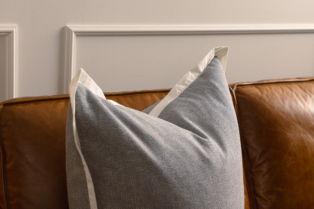 A light blue pillow with a cream trim on an orange leather couch.