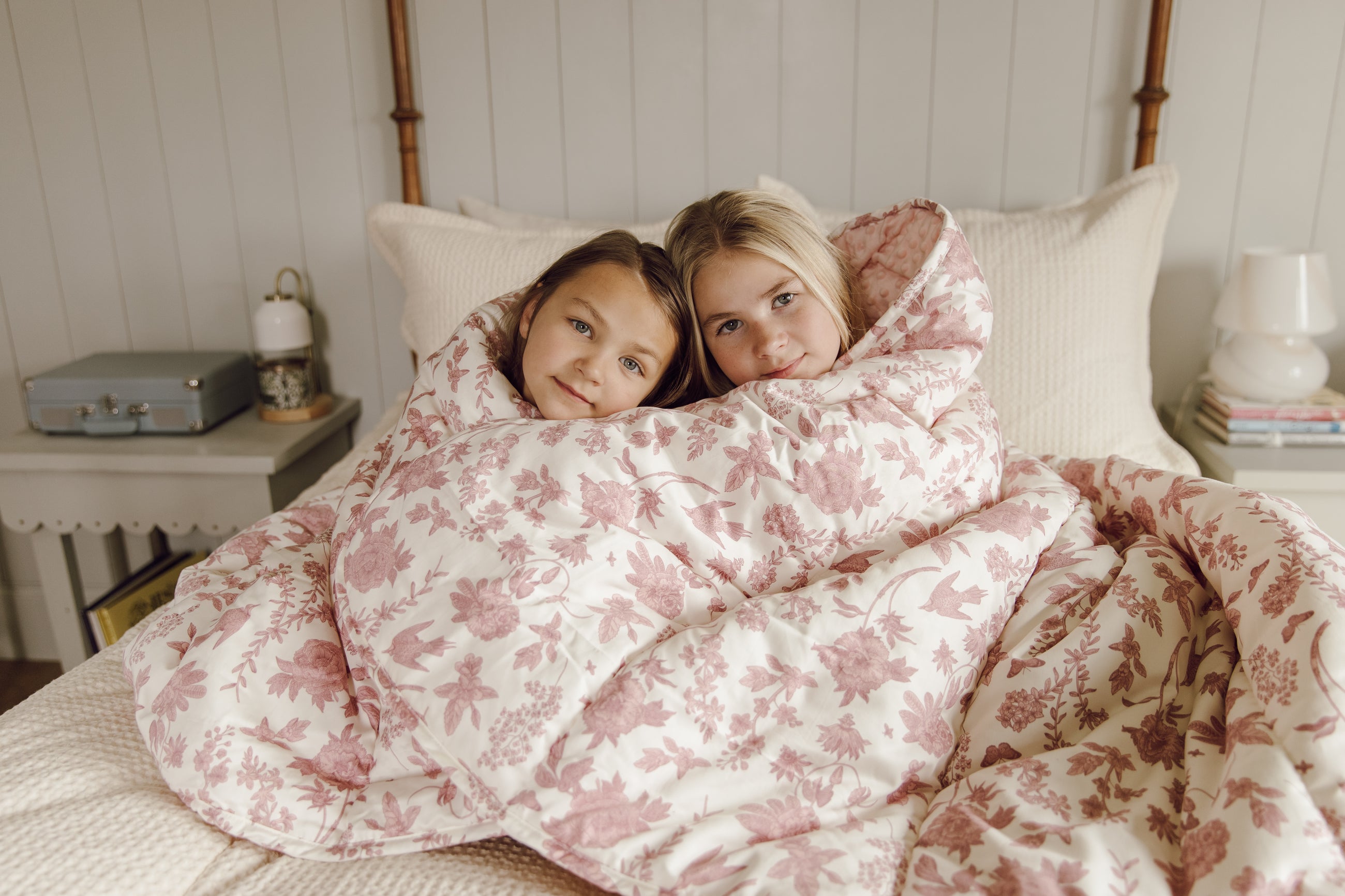 Two girls wrapped in pink and white floral blanket on a bed.