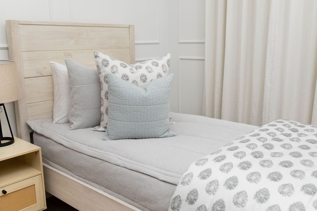 A twin bed made with light gray waffle textured zipper bedding and an assortment of neutral and gray floral pattern throw pillows and blanket.