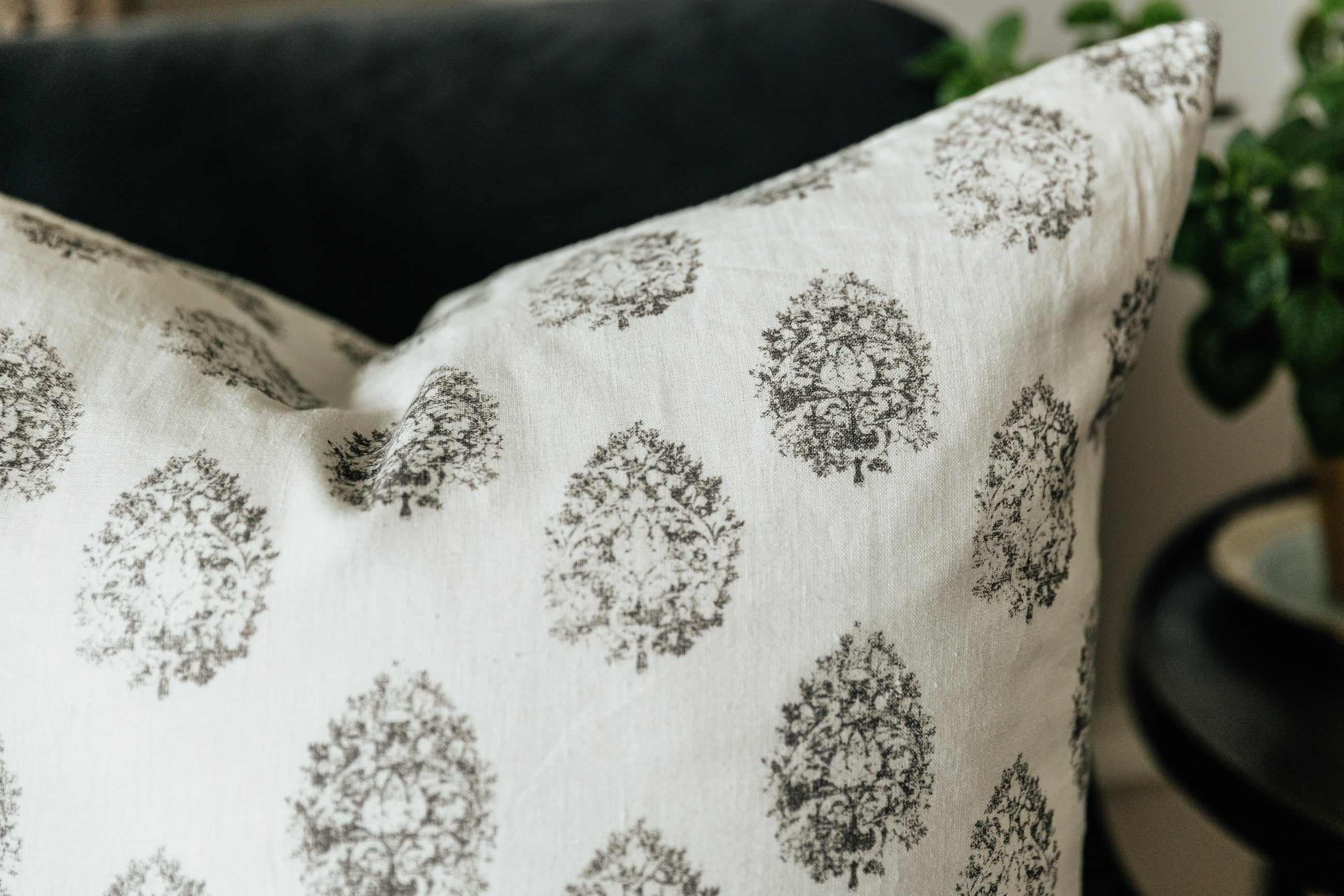 A close up corner view of a charcoal floral print white pillow arranged on a black velvet couch.