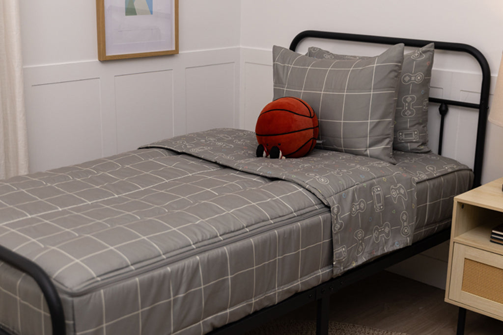 A twin bed made with gaming zipper bedding, a nightstand beside the bed and a stuffed ball propped up against the pillow.