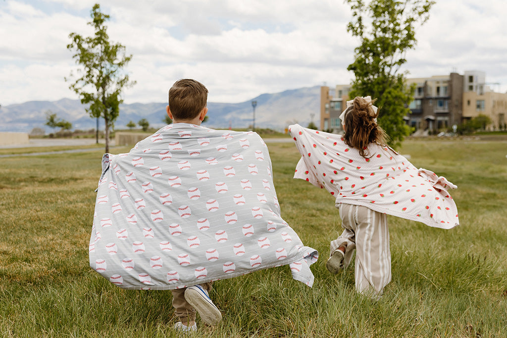 A young boy and girl running through a field of grass wrapped in a little roamer blanket. 