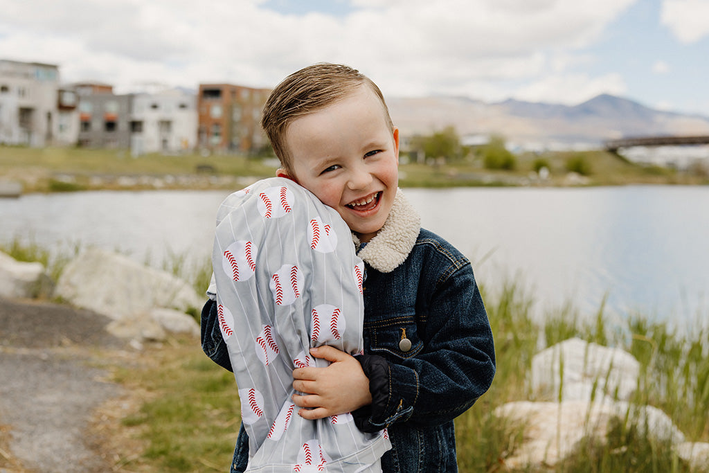 A little boy hugging his baseball print children's blanket while standing outside on a warm summer day.