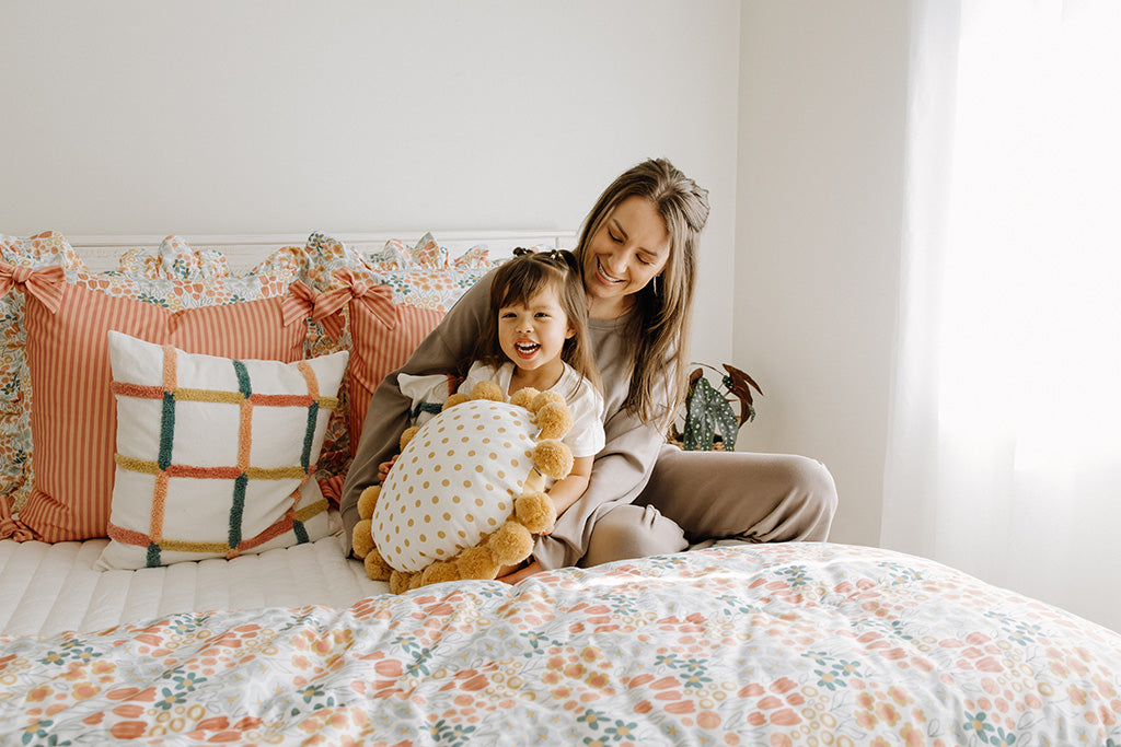 A mother and daughter sitting on white quilted bedding with multi-color and floral accessories. 