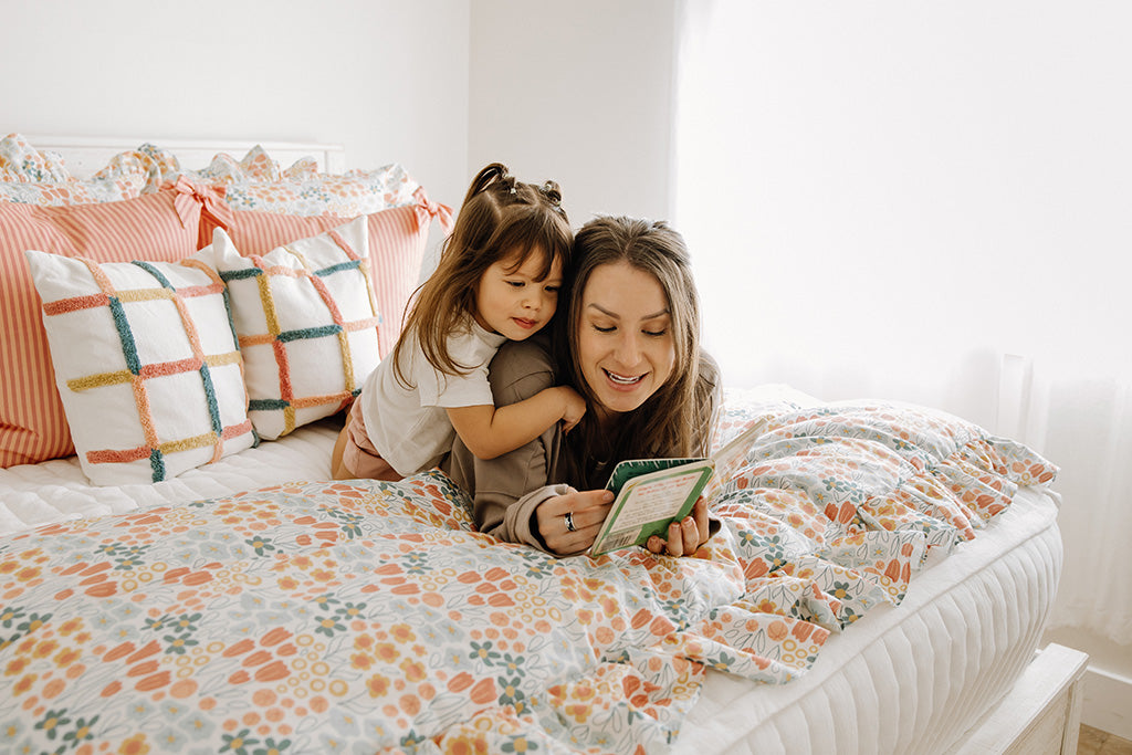 A mom and daughter lying on a white quilted zipper bedding with multi-color and floral accessories reading a book together. 