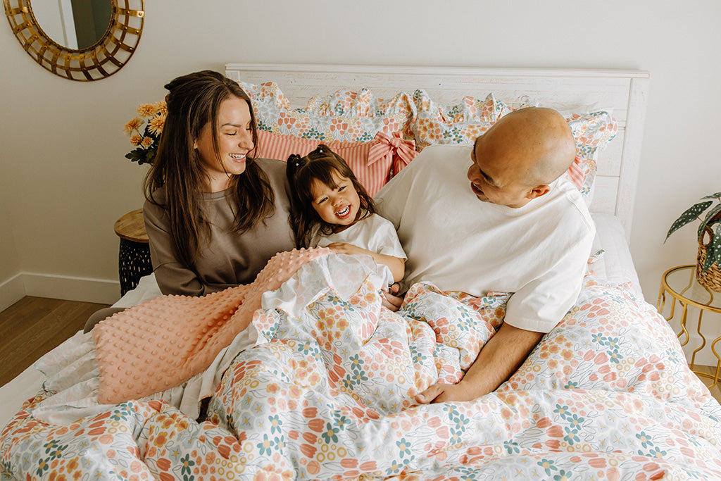 A mom, dad, and daughter snuggling under a multi-color floral blanket. 