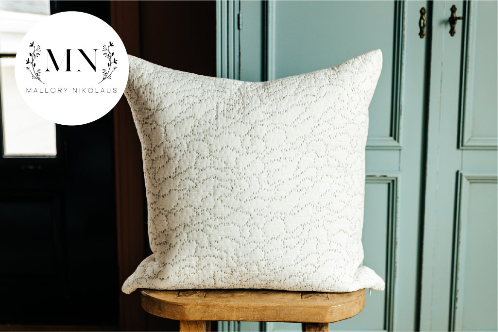 A white euro pillow with intricate green threading arranged on a wood stool in front of a light blue cabinet.