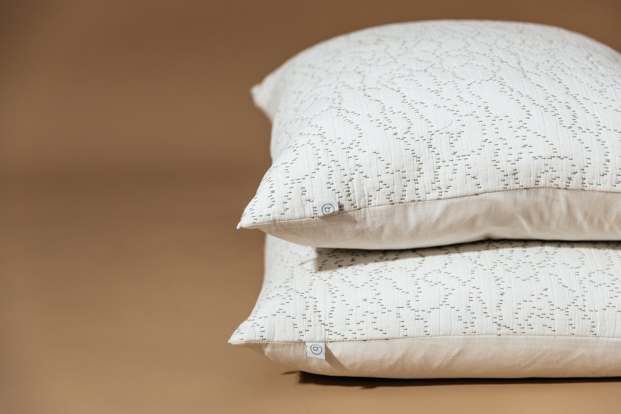 A close up view of two stacked white euro pillows with intricate green threading arranged on a brown background.