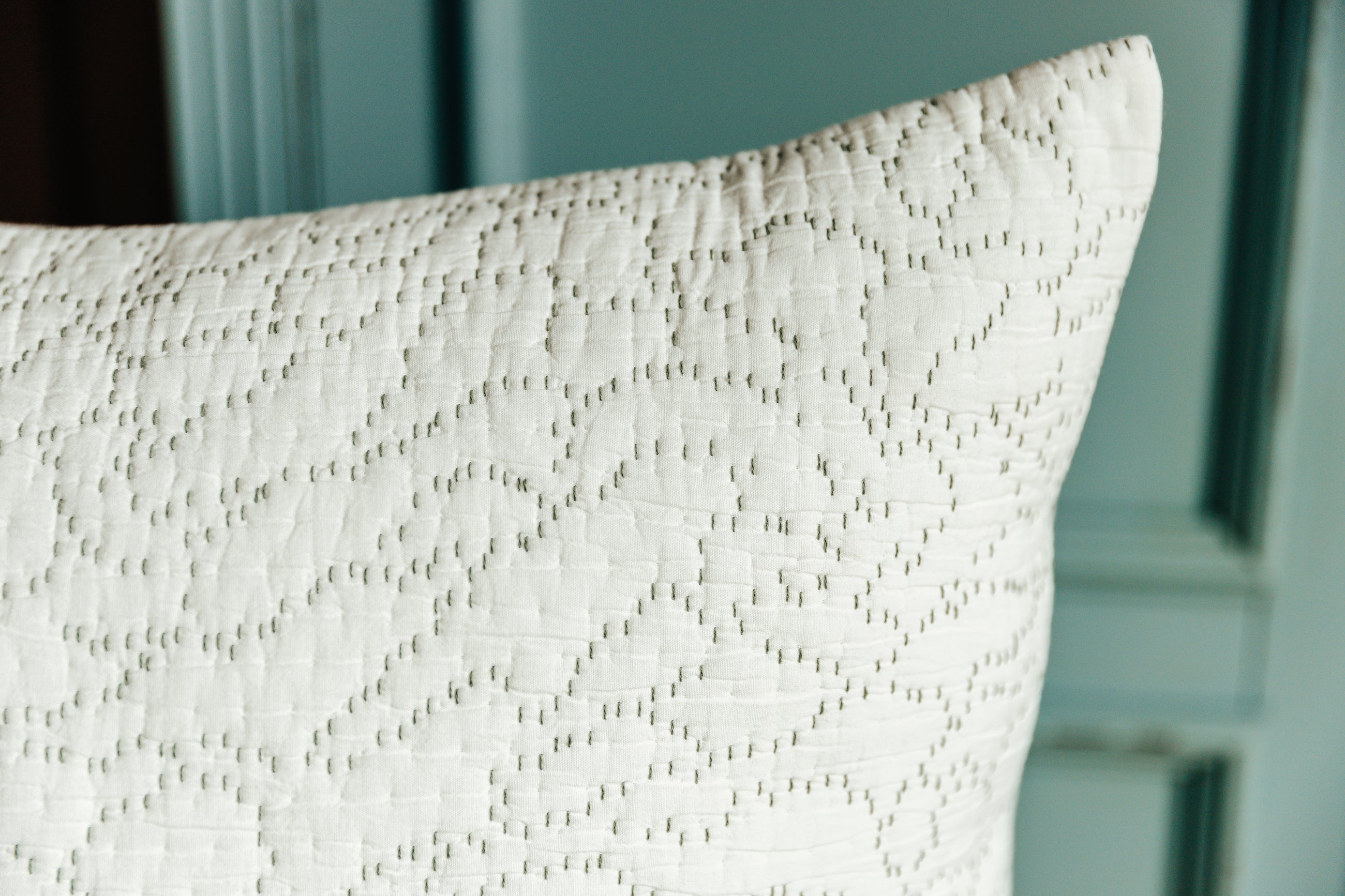 A close up corner view of a white euro pillow with intricate green threading arranged on a wood stool in front of a light blue cabinet.