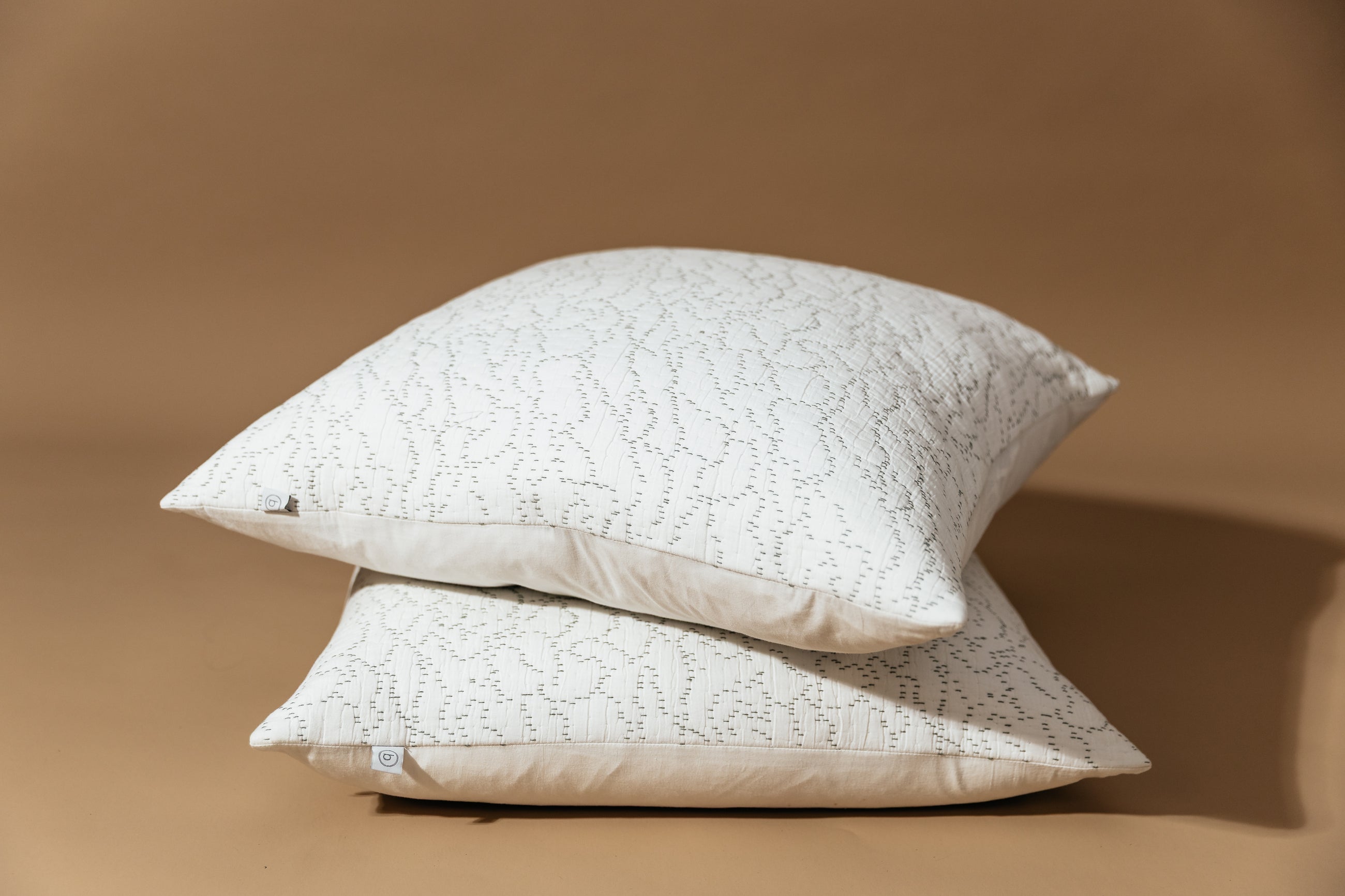 Two stacked white euro pillows with intricate green threading arranged on a brown background.