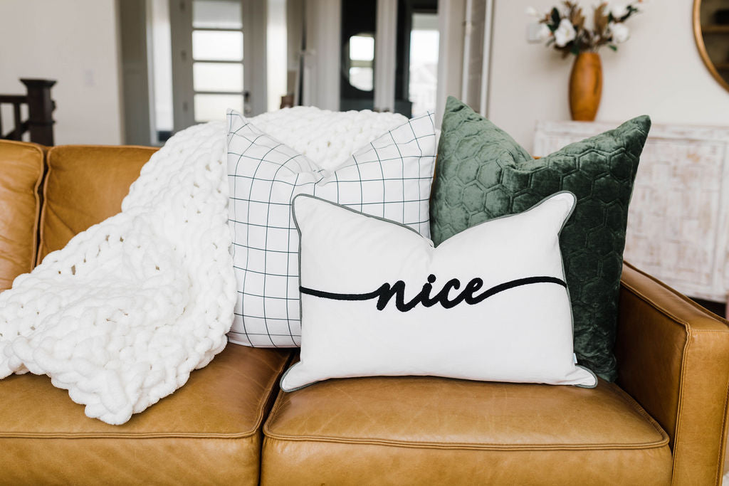 Three white and green velvet pillows arranged on a tan leather couch with a white knit blanket.