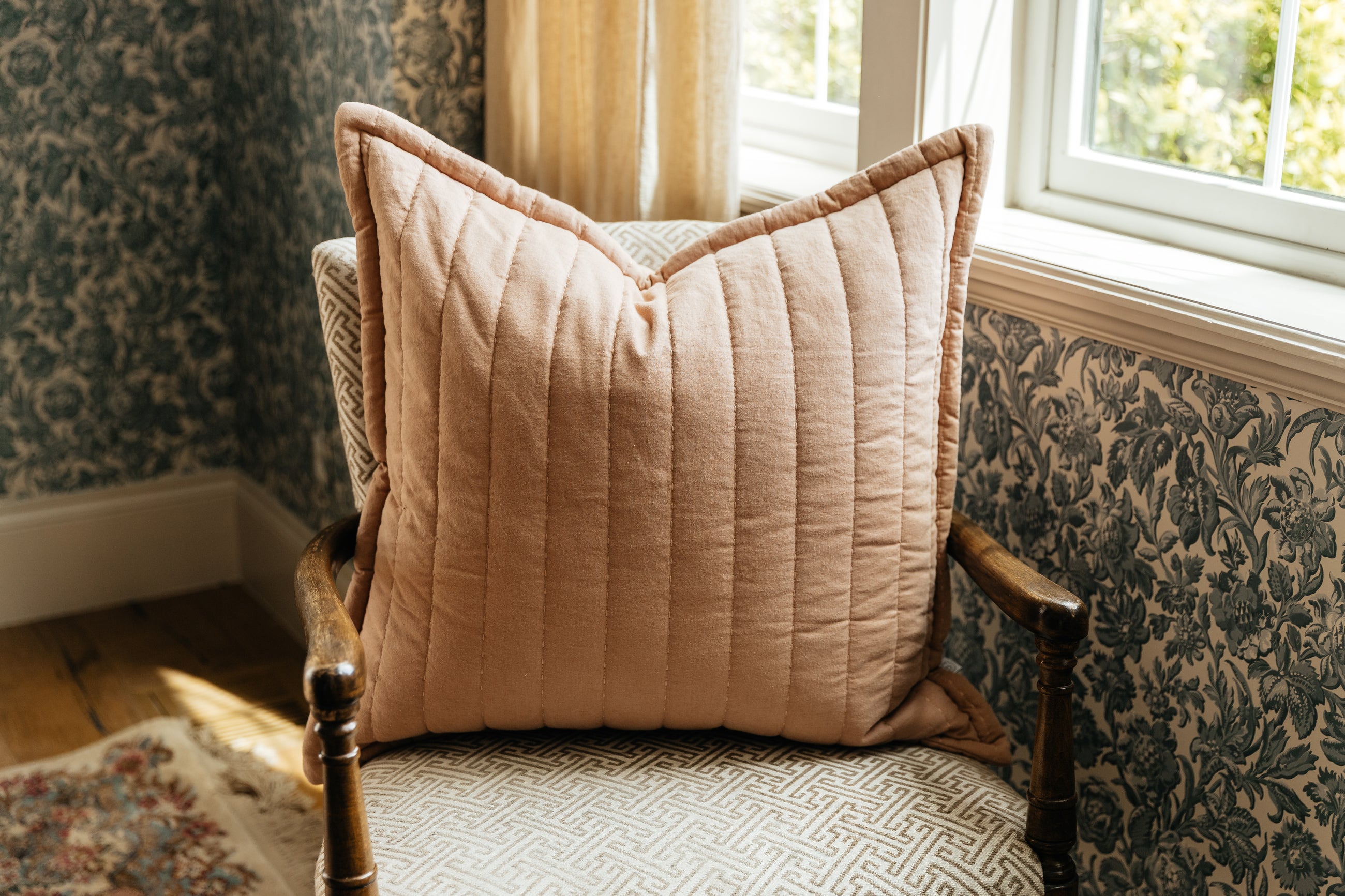 A terracotta quilted pillow on a cream antique chair by a window.