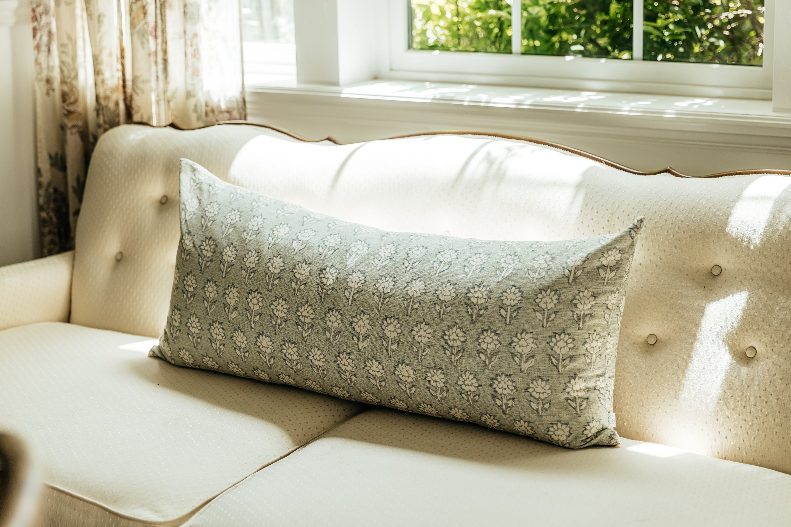 A sage green and blue floral pattern XL lumbar pillow arranged on a cream antique coach by a window.