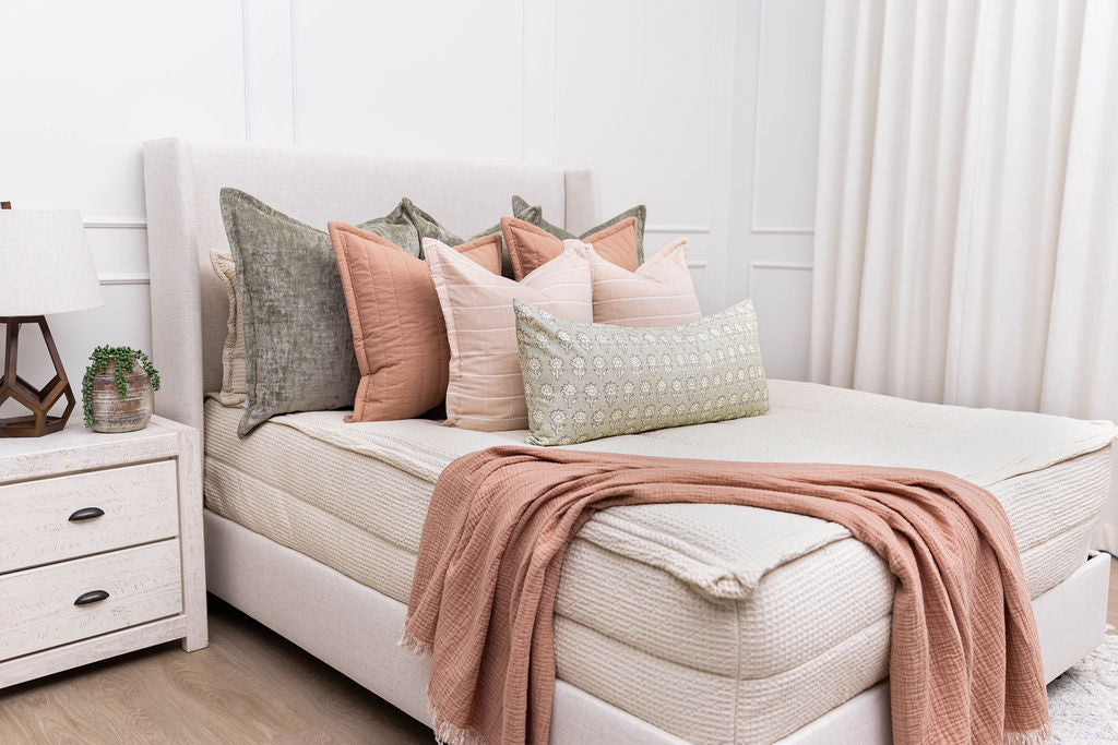 A brightly lit bedroom with a bed made with cream waffle textured zipper bedding and an assortment of green and pink pillows and blanket.