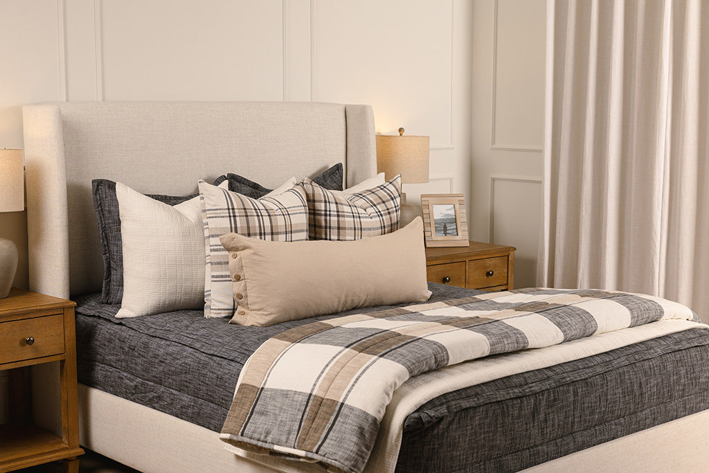 A queen bed made with dark charcoal gray zipper bedding and leather and neutral striped pillows and blanket.
