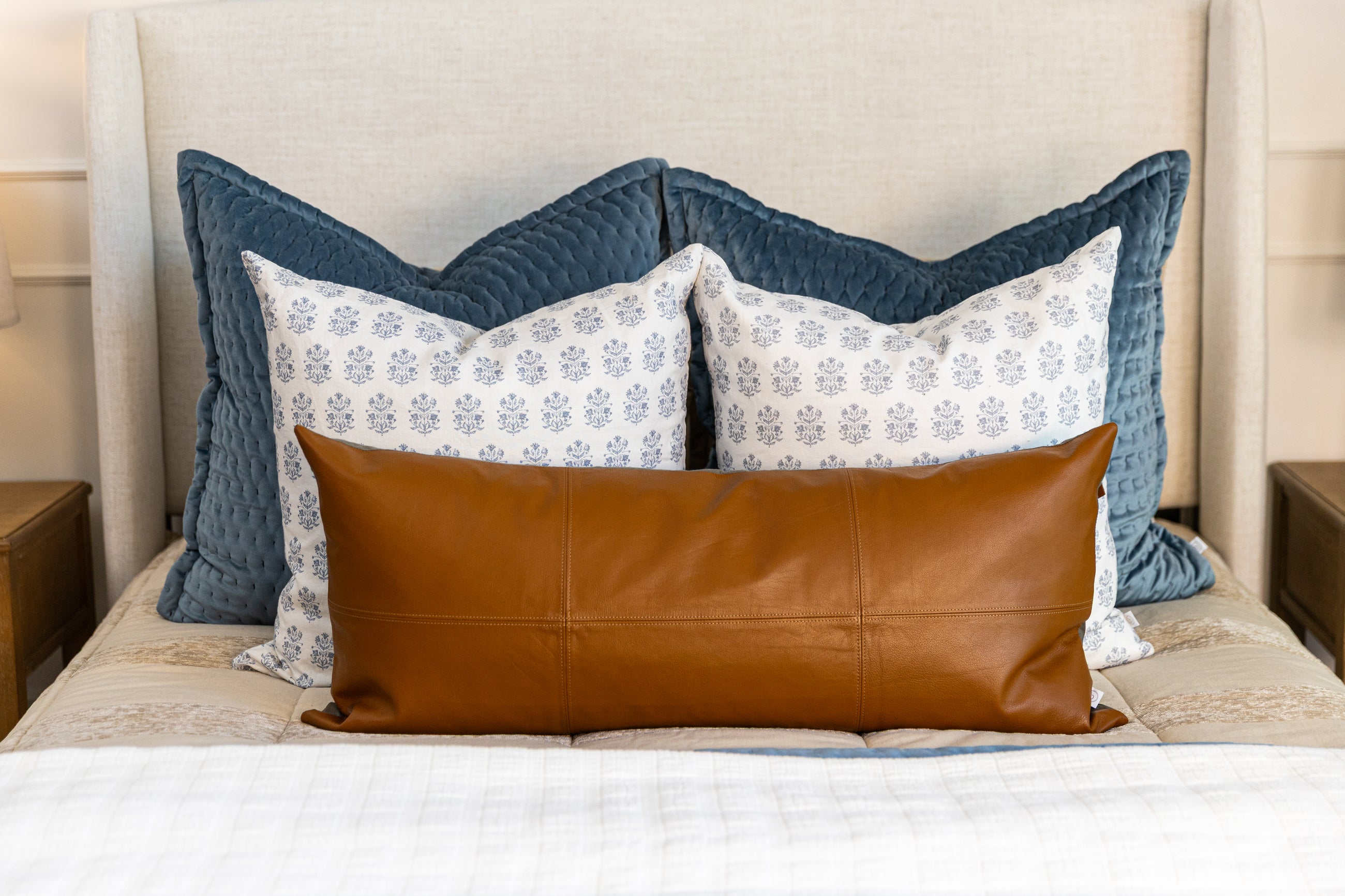 A queen bed made with tan zipper bedding and an arrangement of velvet, floral pattern, and leather pillows.