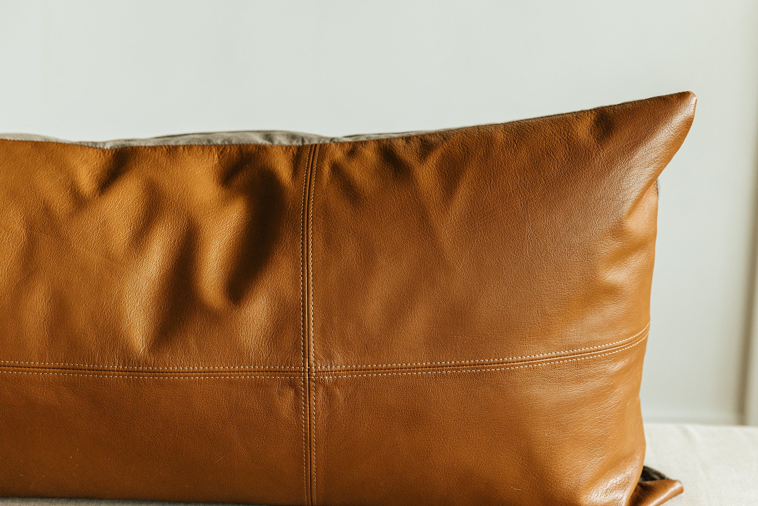 A close up view of a neutral leather XL lumbar pillow with stitching arranged on a cream bench.