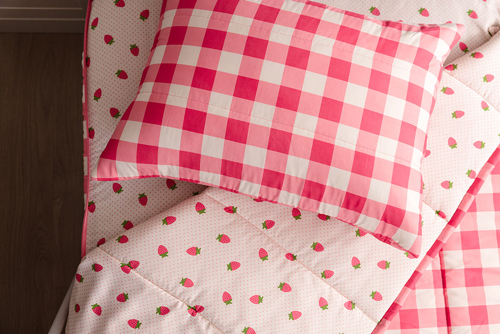 Pink gingham and strawberry printed kids zipper bedding with the top unzipped and one side pulled back to reveal the inside lining, the pillow tossed on the top of the bed.