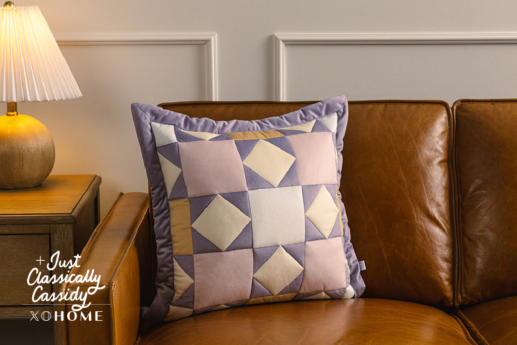 A purple, pink, cream, and yellow quilted pillow on an orange leather couch.