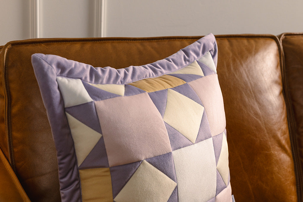 A purple, pink, cream, and yellow quilted pillow on an orange leather couch.