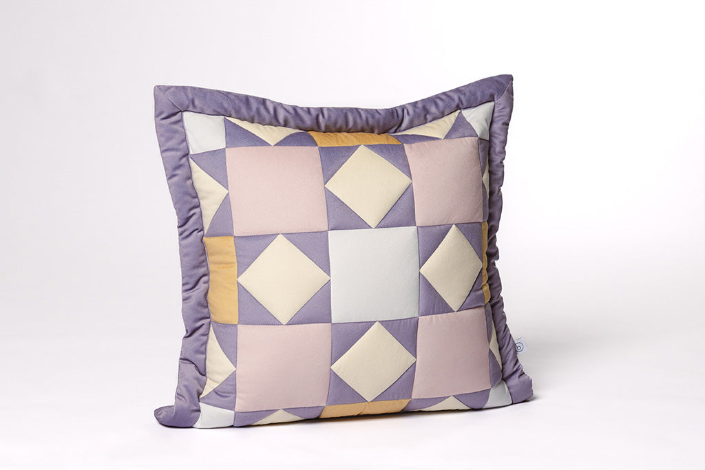 A purple, pink, cream, and yellow quilted pillow on a white backdrop.
