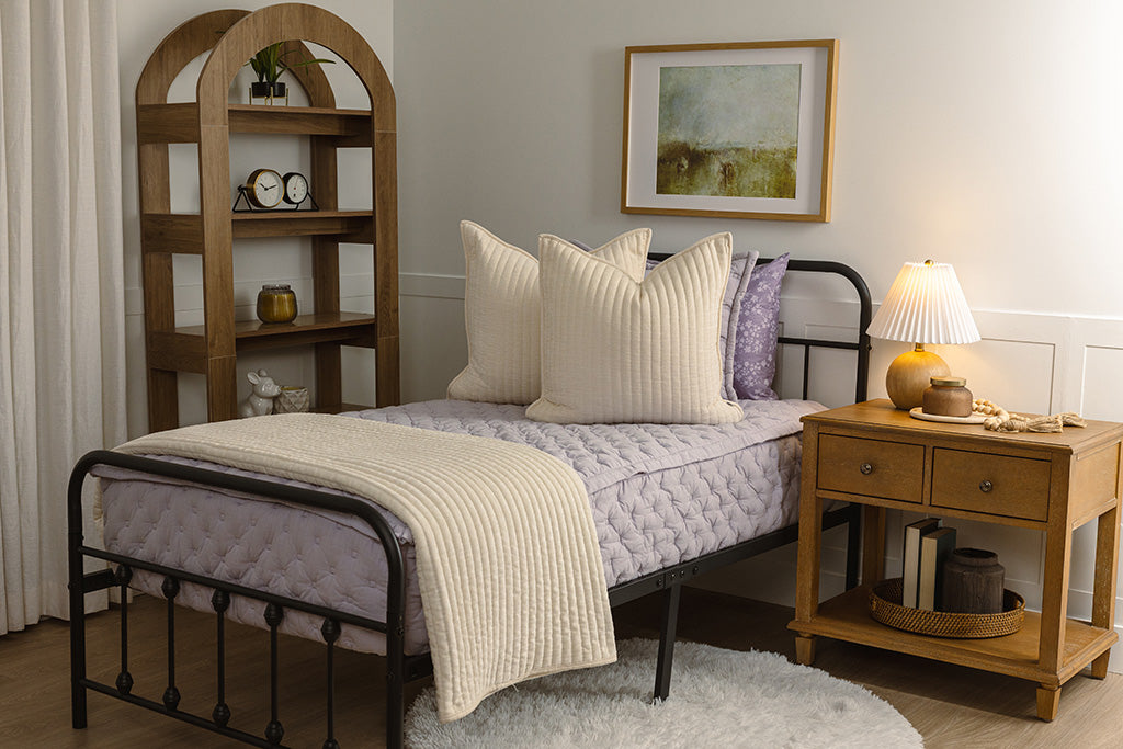 A twin bed with purple puffed bedding with cream accessories. 