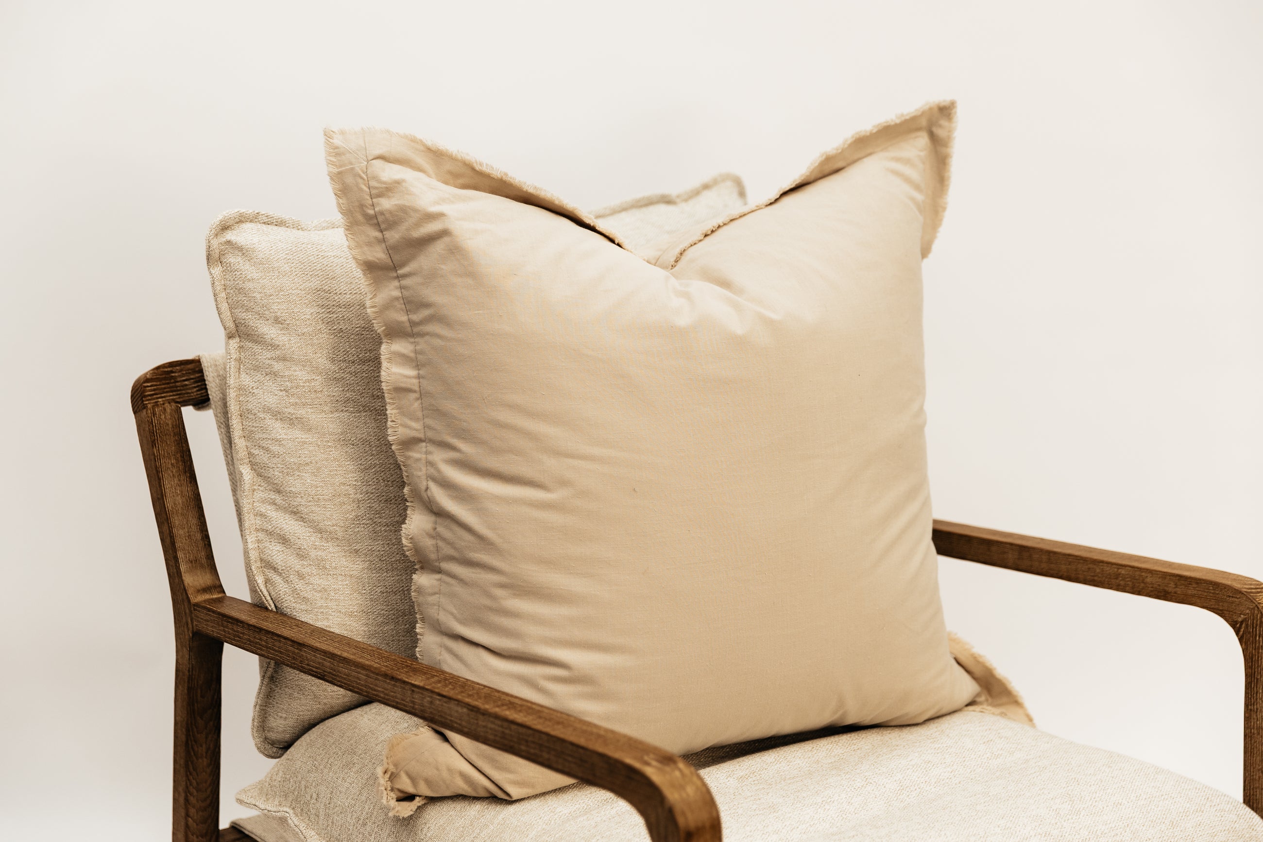 A neutral large tan pillow with frayed edges arranged on a cream fabric chair.