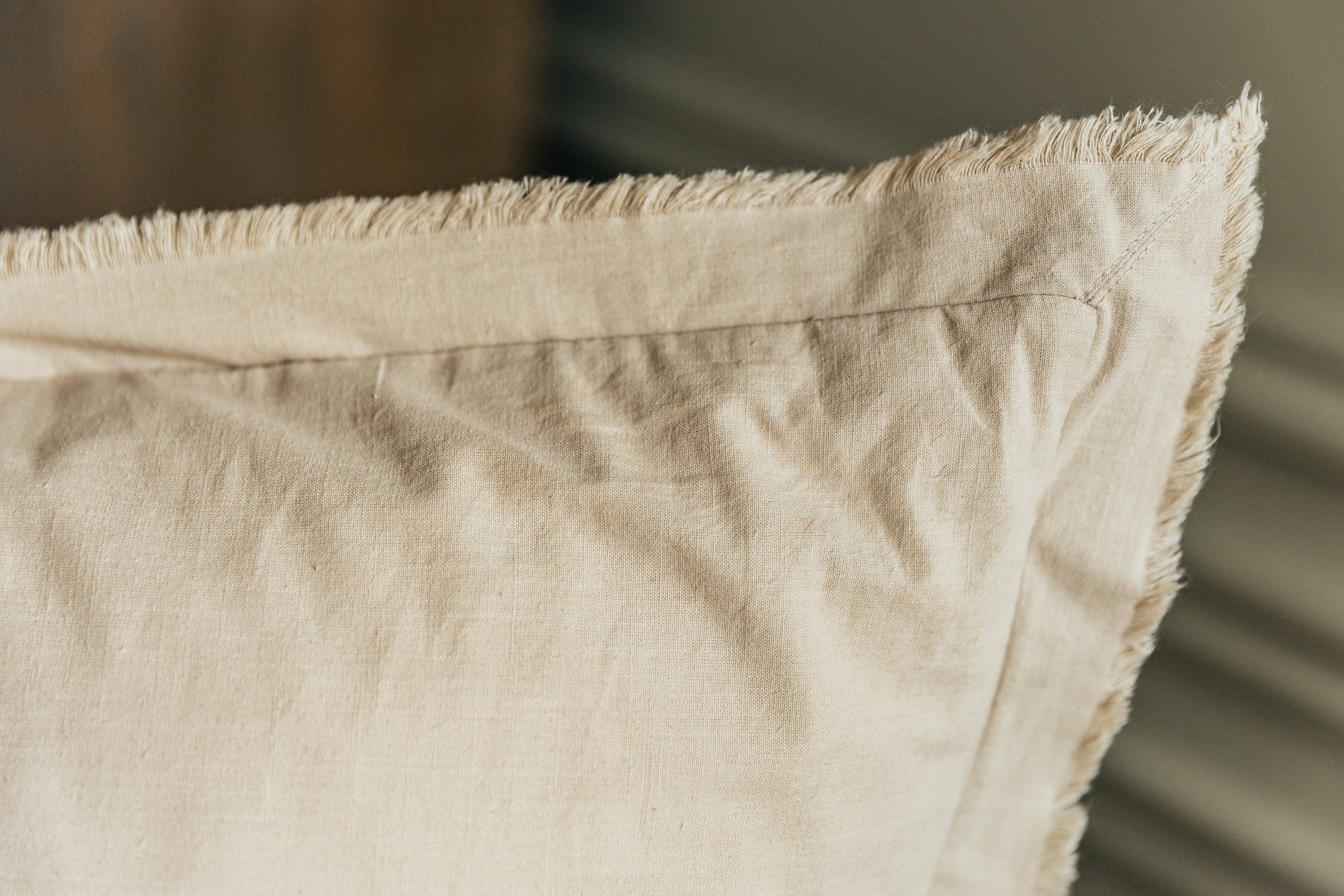 A close up corner view of a neutral large tan pillow with frayed edges.
