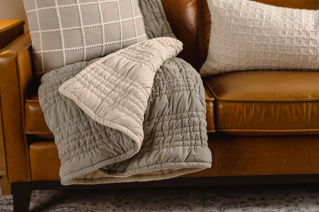 A sage green and cream blanket on an orange leather couch with cream and tan pillows.