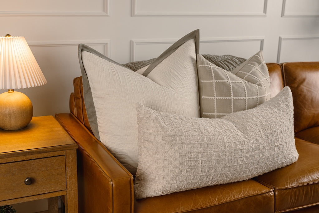 A cream textured and green velvet trim pillow and tan and a cream trimmed grid pillow, with a cream knitted lumbar pillow on a leather couch.