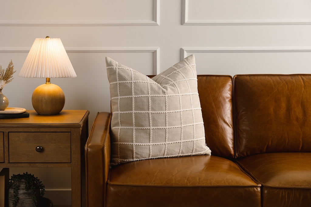 A tan and a cream trimmed grid pattern pillow on a leather couch.