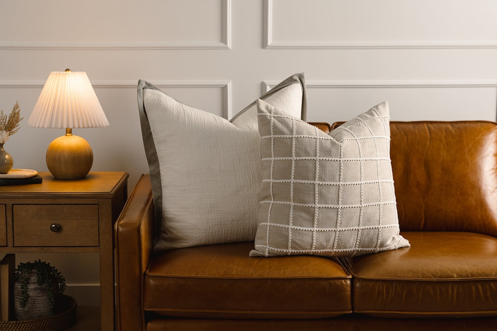 A cream textured and green velvet trim pillow and tan and a cream trimmed grid pillow on a leather couch.