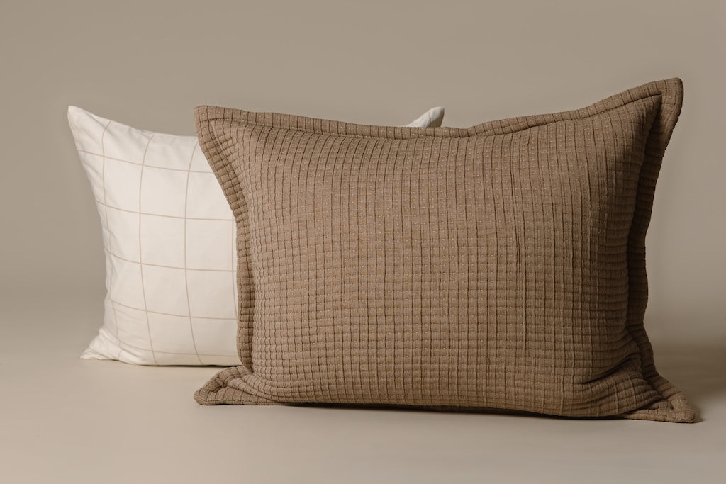 A cream and tan grid pillow and a light brown knitted sham on a cream backdrop.