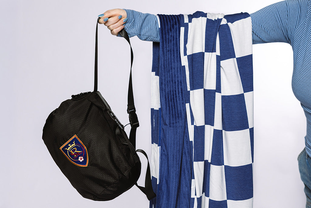 A woman with a blue and white check blanket draped over her arm and holding a black bag on a white backdrop.