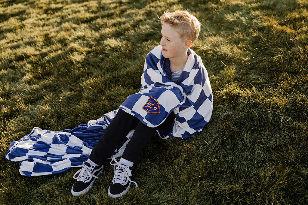 A young boy wrapped in a blue and white check blanket sitting in a green grass field. 