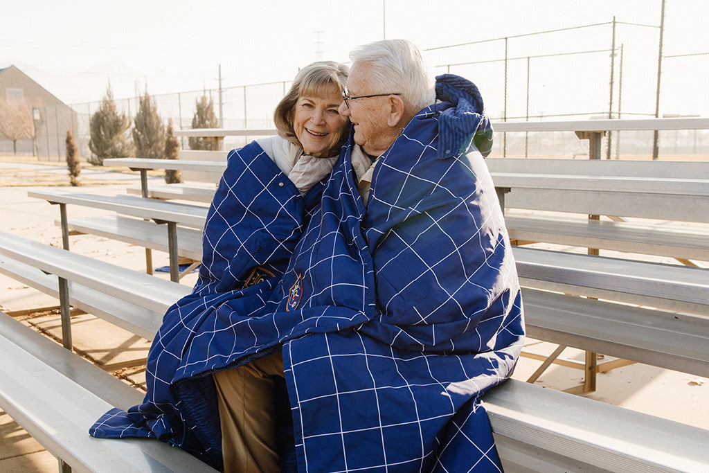 A couple wrapped in a blue blanket with white grid stripe blanket, outdoors in bleachers. 
