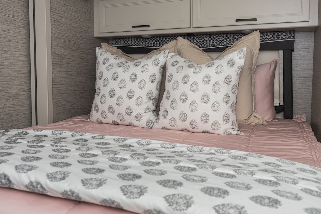 A bed made in an RV with pink zipper bedding and an assortment of cream, white, and grey floral pillows and blanket.