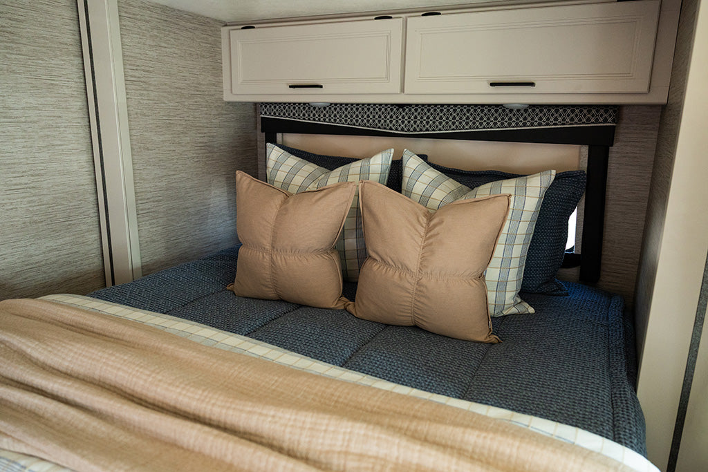 A bed made in an RV with dusty blue knitted zipper bedding and an assortment of tan, white and blue plaid pillows and blankets.