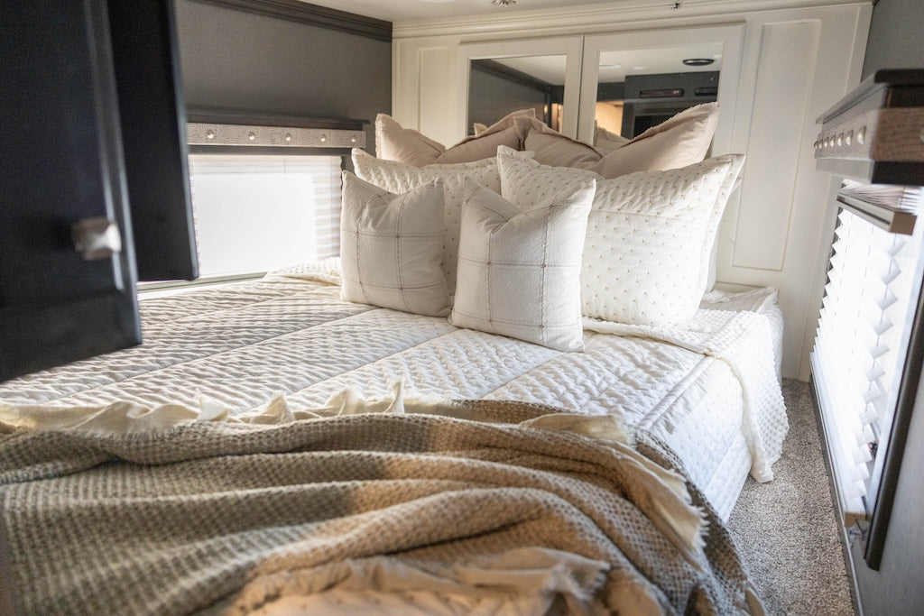 A bed in an RV made with cream embroidered zipper bedding and neutral pattern pillows and a throw blanket.