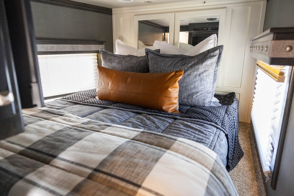 A bed made in an RV with dark charcoal gray zipper bedding and an assortment of neutral and plaid throw pillows and blanket.