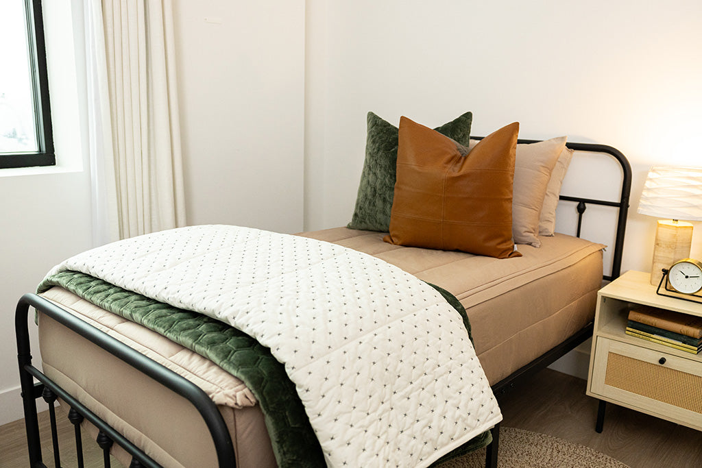 A close up view of a twin bed made with tan zipper bedding and green and neutral accessories. 