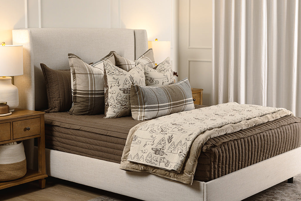 A cream and brown quilted holiday blanket  on brown quilted bedding with tan, brown, and white accessories.