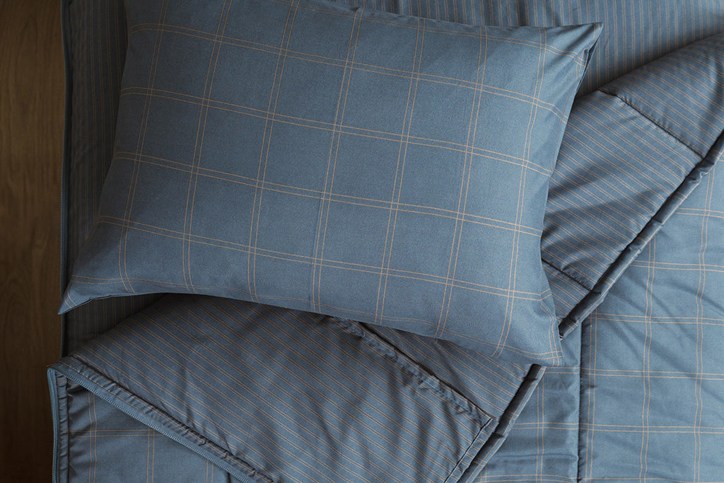 Slate blue grid printed zipper bedding with the top unzipped and one side pulled back to reveal the inside lining, the pillow tossed on top of the bed.