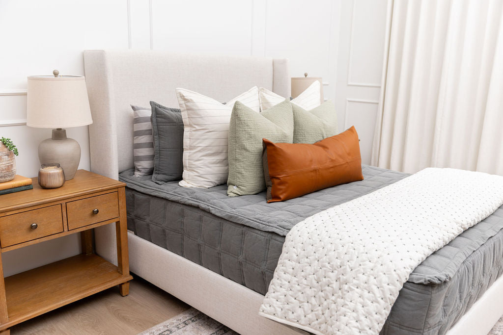 A bed made with gray zipper bedding with an assortment of cream, sage green and leather pillows and blanket.
