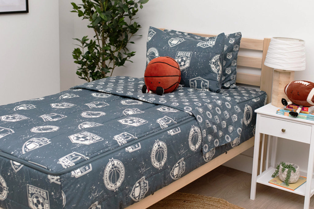 A twin bed made with blue sports themed kids zipper bedding and a nightstand beside it holding a lamp and a stuffed football